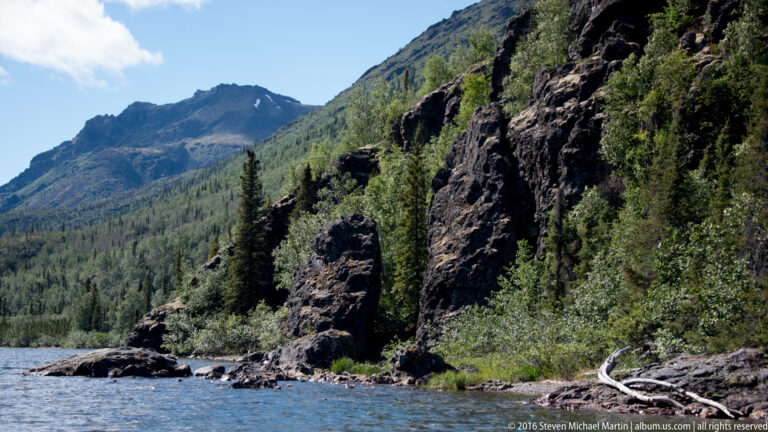 Fractured dark cliffs and trees rise above a rocky point at the lake’s edge.