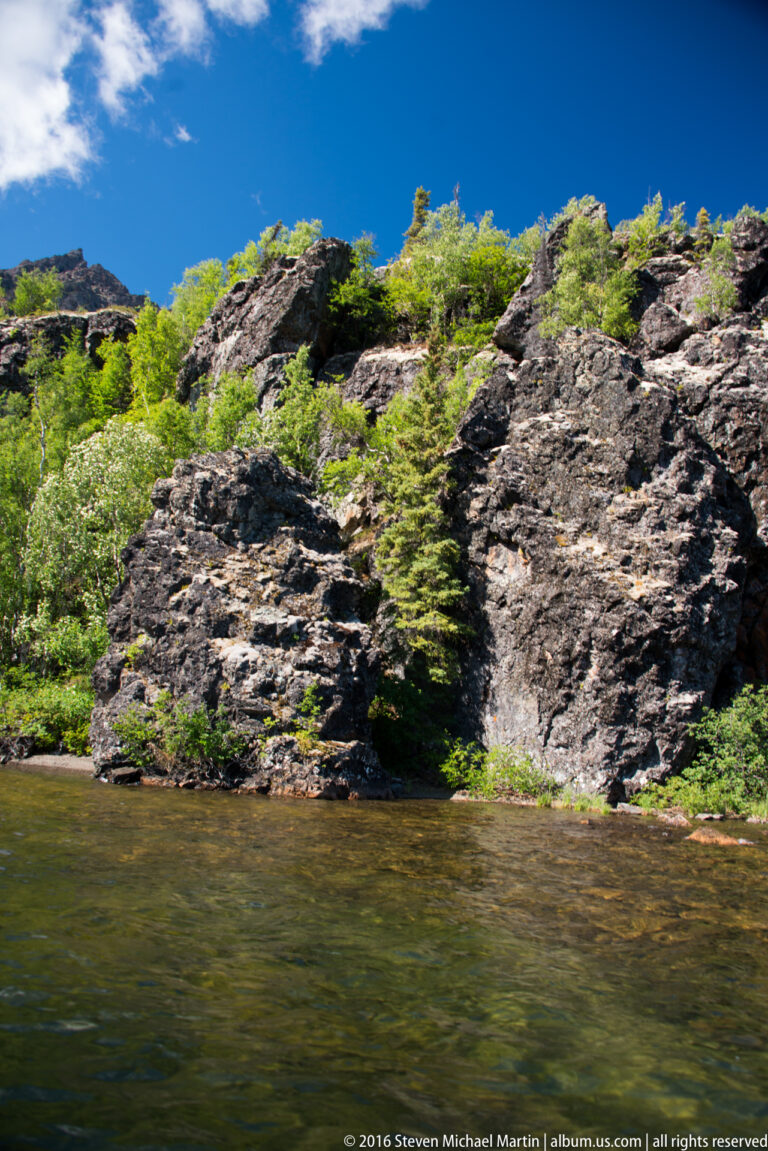 Dark rocky ledges with shrubs meet the water beneath a bright blue sky.