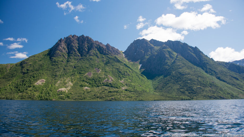 Jagged green mountains rise above blue lake water beneath a bright cloud.