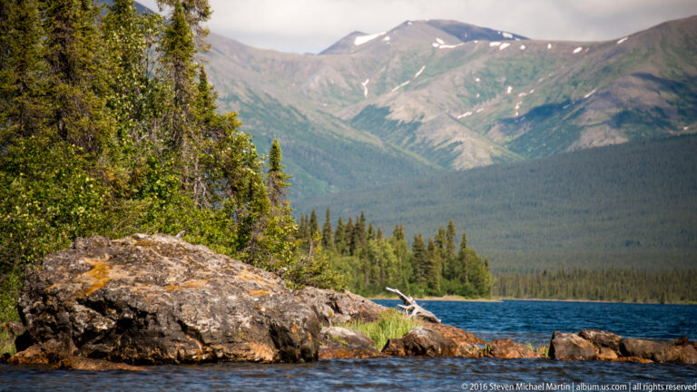 Rocky shore overlooks a forested valley and distant mountains with small snow patches.