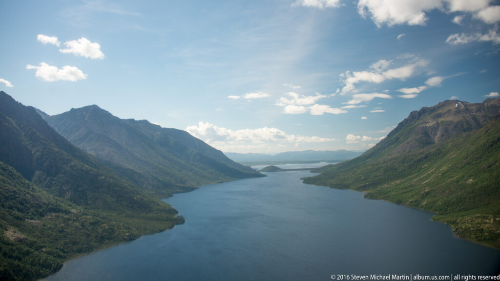 A long lake narrows between steep forested mountains toward a distant opening under scattered clouds.
