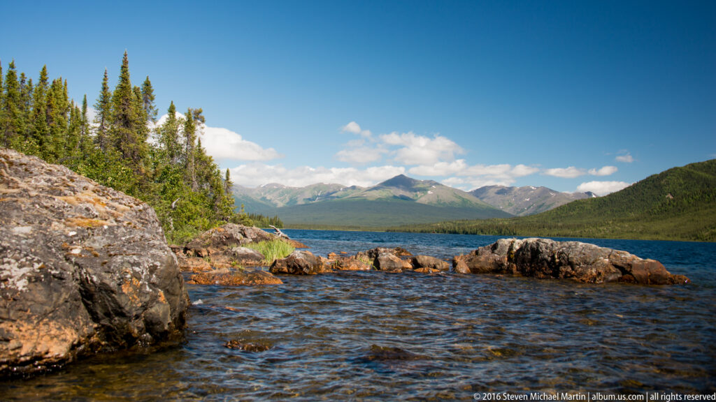 Rock ledges line the foreground of a lake with forested slopes and distant peaks.