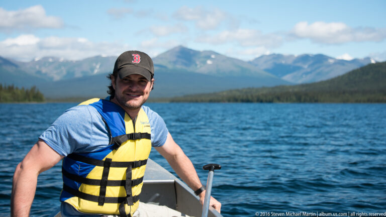 A man in a yellow-and-blue life jacket sits in a small boat on blue water, with forest and mountains behind him.