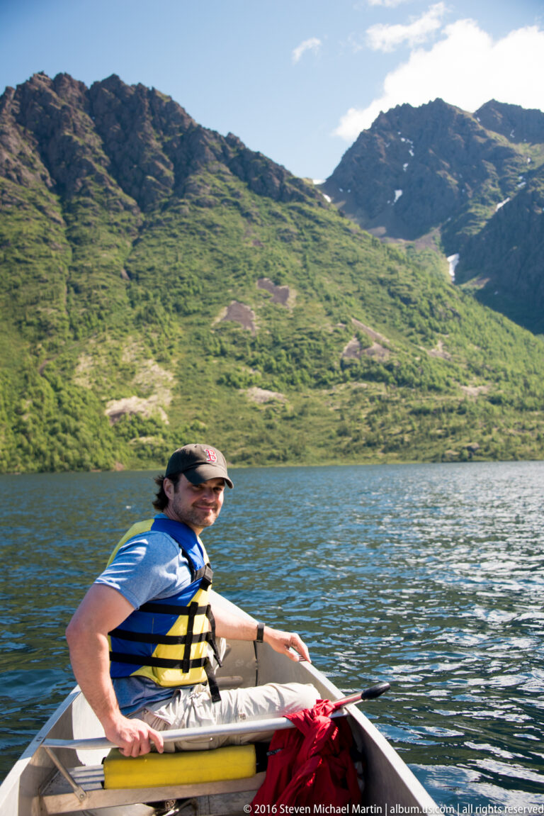 A man wearing a life jacket holds a paddle in a small boat on a lake beneath green mountain slopes.