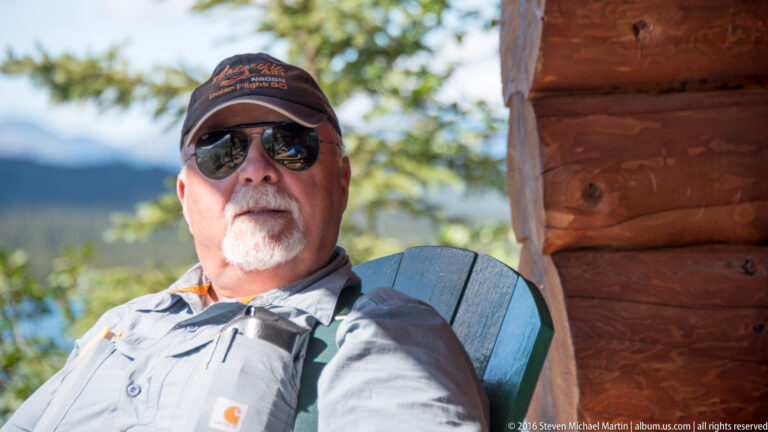 A seated man in a cap and mirrored sunglasses rests beside a log wall, with trees and mountains behind him.