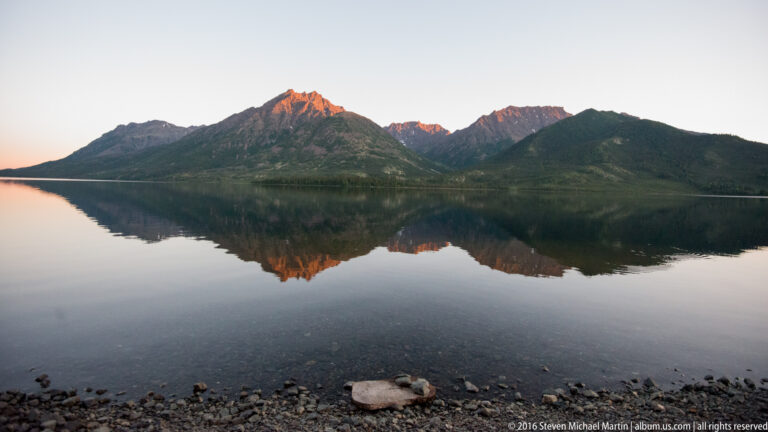 Orange-lit mountain peaks and a dark shoreline reflect across a calm lake at dusk.