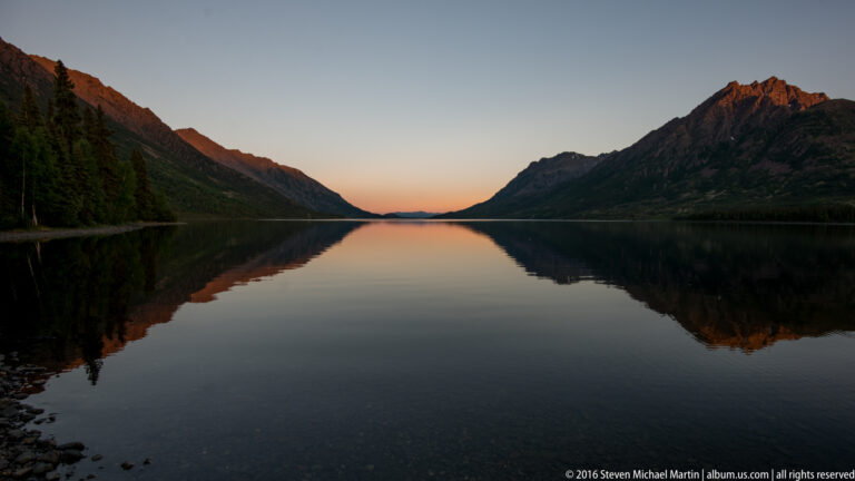 A long lake stretches toward a warm twilight horizon between dark mountain slopes, with the scene reflected in still water.