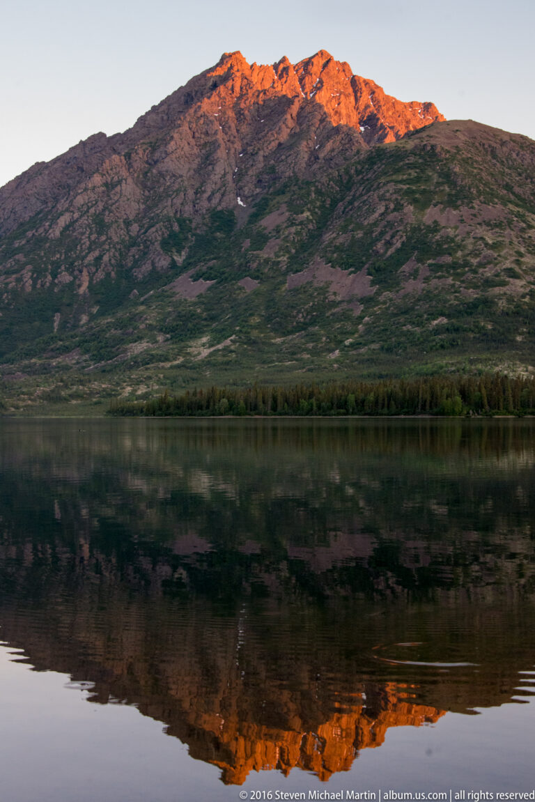 Orange alpenglow touches rocky summits above darker slopes, with the mountain reflected in a still lake.