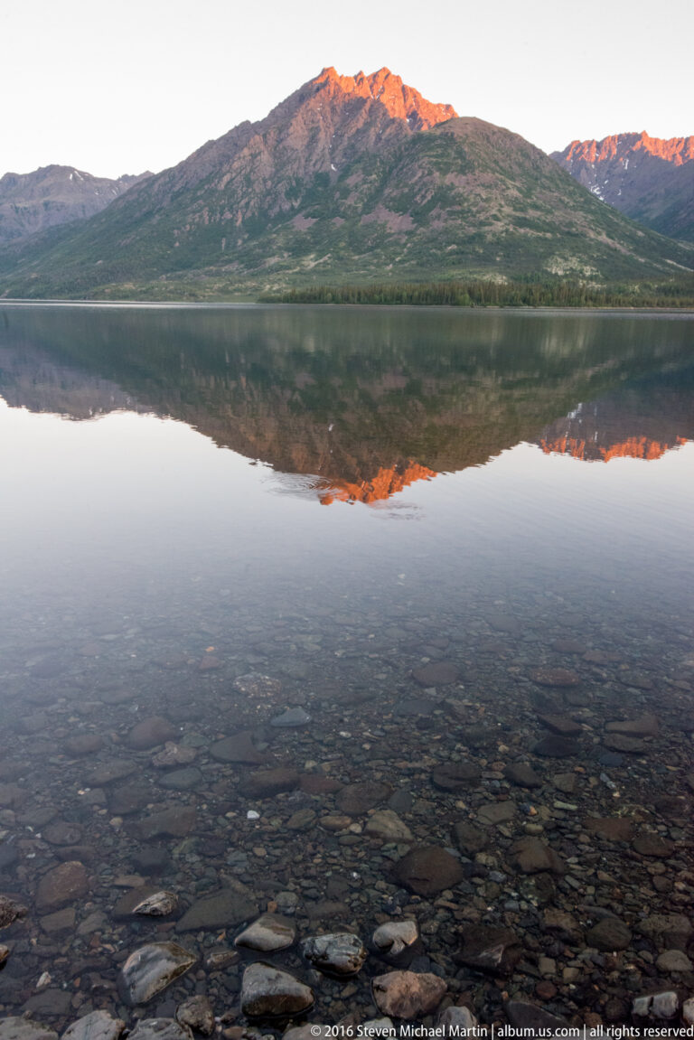Submerged stones show through clear lake shallows beneath the reflection of an orange-lit mountain ridge.