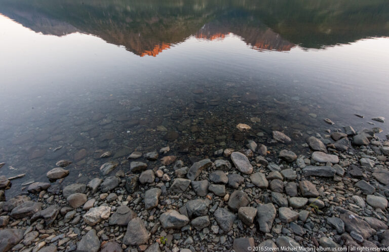 Rounded stones beneath shallow clear water fill the foreground, below reflected mountains and sky.