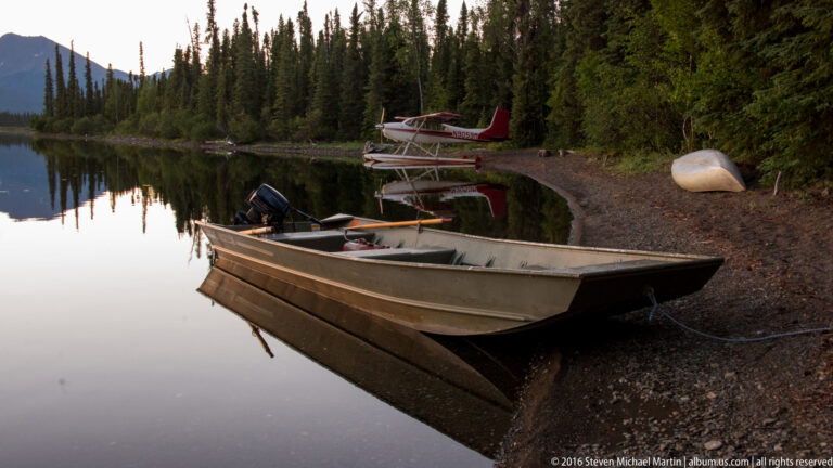 An aluminum motorboat rests at a wooded lake landing, with a floatplane across the cove and a canoe on shore.
