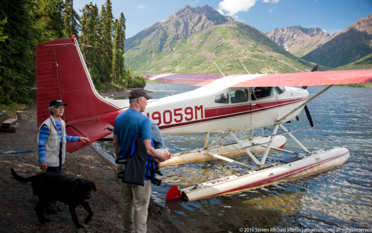 A red-and-white floatplane sits on the shore of a mountain lake beside two people and a black dog.