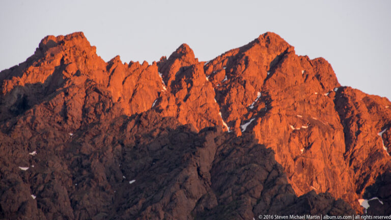 Jagged rocky mountain peaks glow orange in low sunlight above shadowed lower slopes.