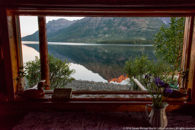A cabin window frames a mountain reflected in the lake, with purple flowers and small objects on the sill.