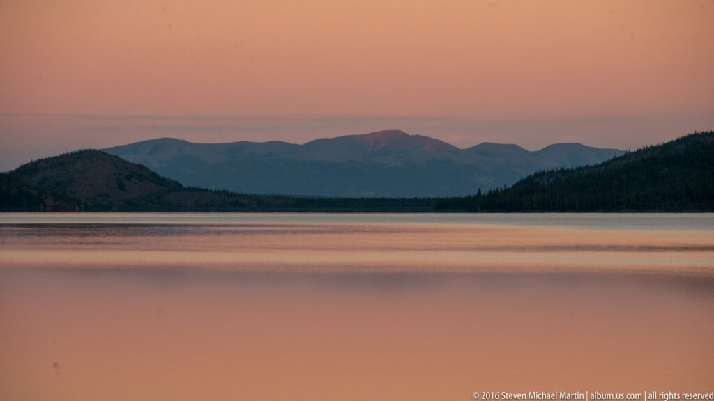 A pink-orange twilight sky and layered dark mountains reflect across a smooth lake.