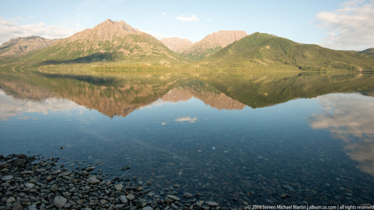 Green and rocky mountain slopes reflect in a clear lake, with rounded stones visible beneath the foreground water.