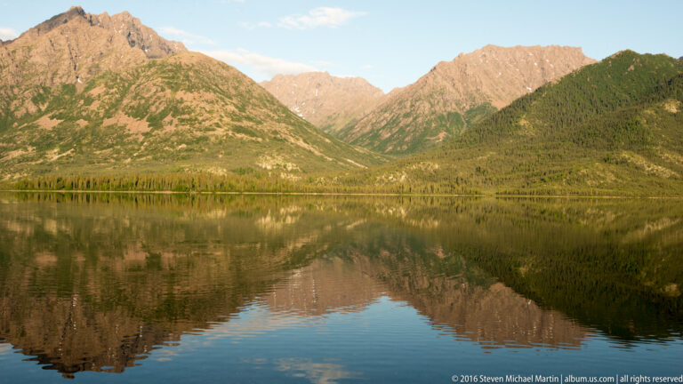 A broad mountain valley and forested slopes reflect in calm lake water beneath a blue sky.