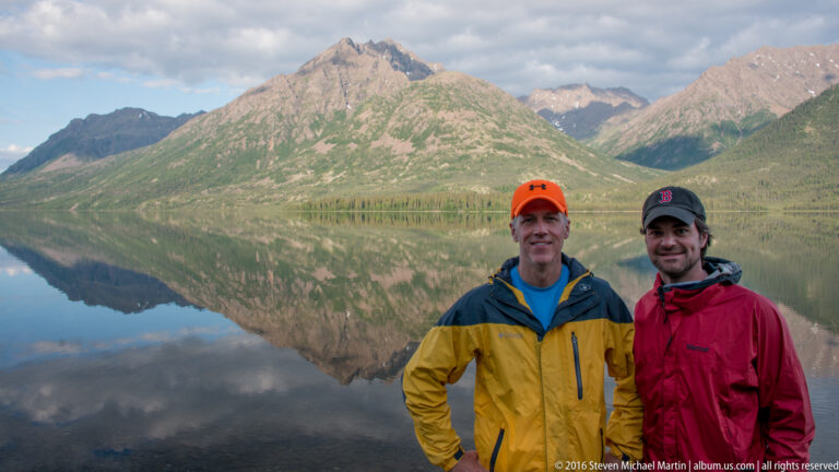 Two men in yellow and red jackets stand on a rocky shore before a reflected mountain.