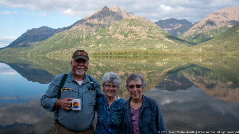An older man holding a mug poses with two women at a rocky lakeshore, with a mountain reflection behind them.