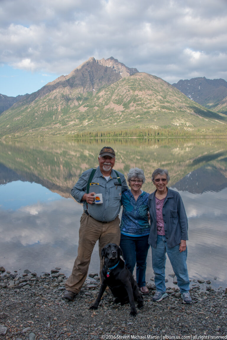 Three adults and a black dog stand on a rocky lakeshore beneath a mountain reflected in the water.