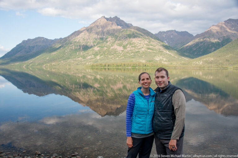 A woman in turquoise and a man in a dark vest stand at the rocky shore before a reflected mountain.