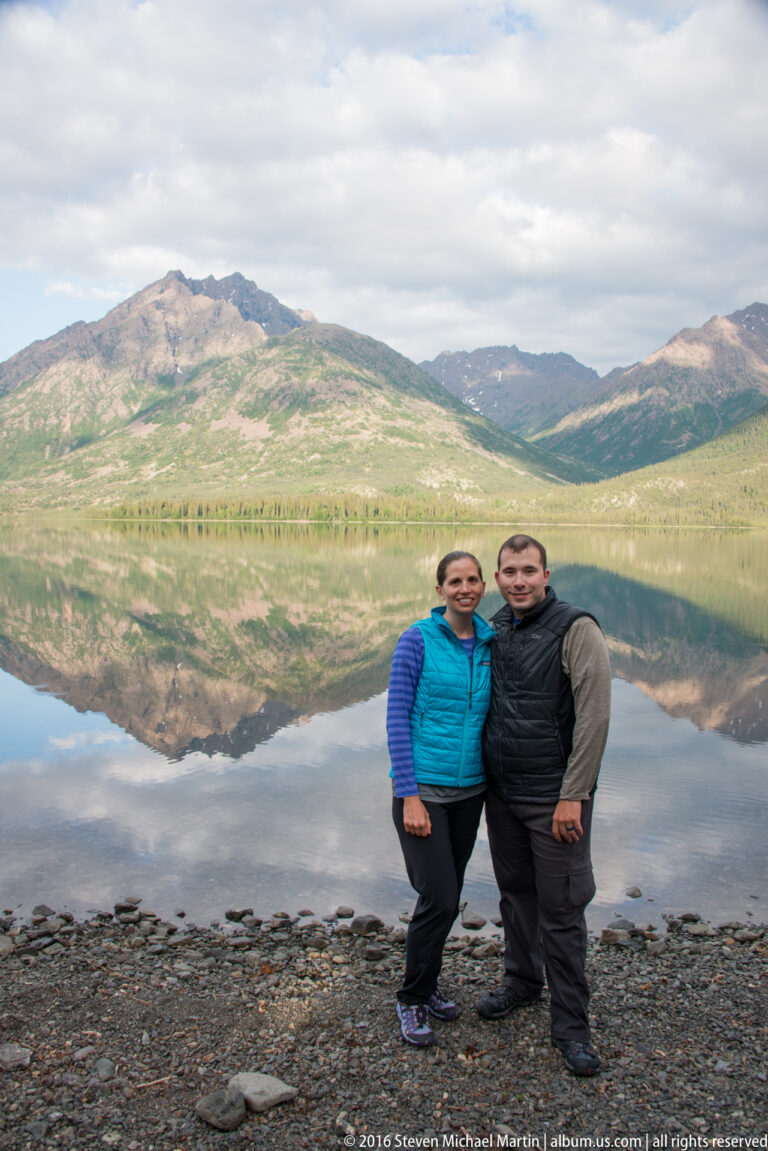 Two adults stand full length on a rocky lakeshore, with a long mountain ridge reflected across the water.