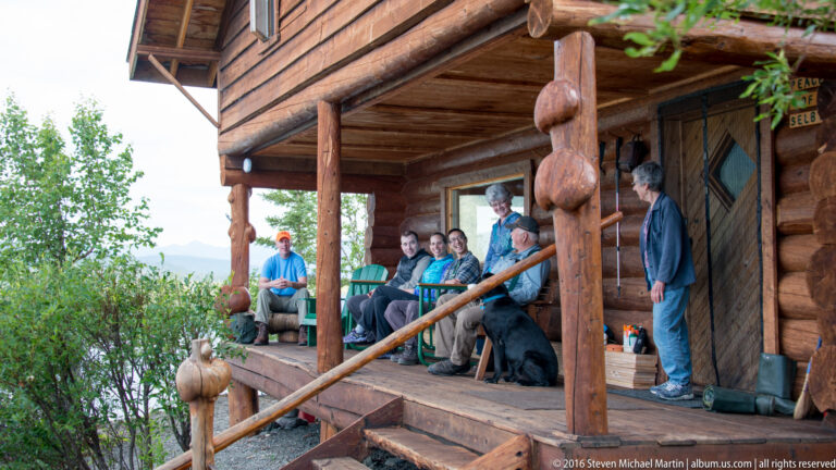 Adults gather on a log cabin porch with a black dog, framed by heavy timber posts and log walls.