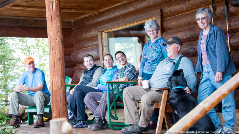 Six adults sit and stand on a log porch beside a black dog, with a cabin window and heavy beam behind them.