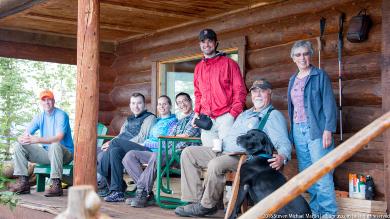 A man in a red jacket stands behind seated companions on a log porch, with two women and a black dog nearby.