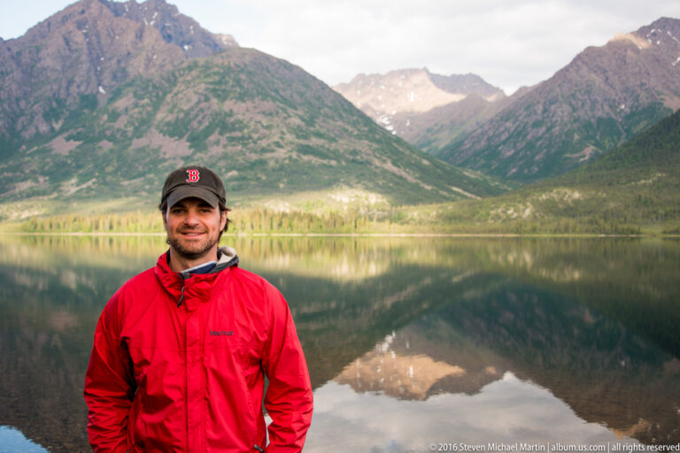 A man in a red jacket and cap stands before a mountain reflected in the lake.