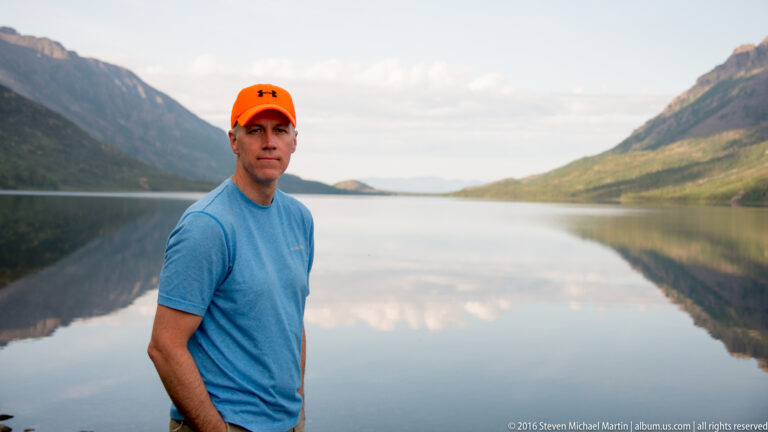 A man in an orange cap and blue shirt stands beside a calm lake, with mountains behind him.