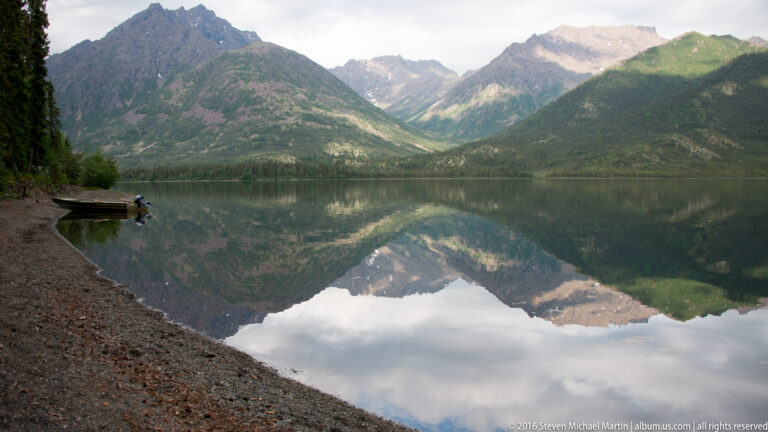 Clouds and mountain slopes reflect across a lake viewed from a pebbled shore, with a small boat at left.
