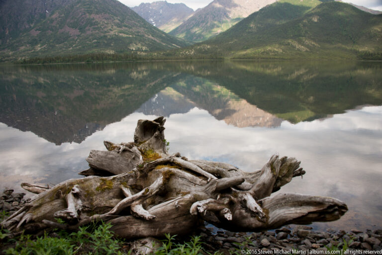 Weathered driftwood lies among stones at the water’s edge, with reflected mountains and clouds behind it.