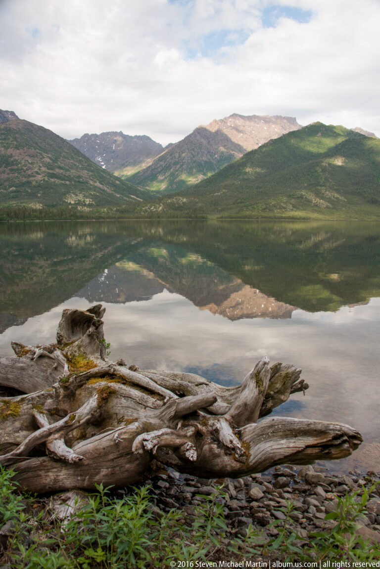 A long driftwood trunk crosses a stony shore beside a lake reflecting green mountain slopes and clouds.