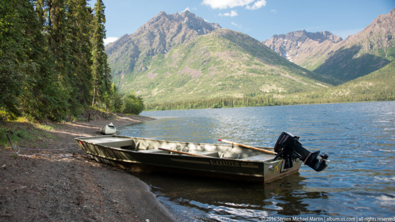 A long motorboat with an outboard rests beside a pebbled shore, with mountains across the lake.