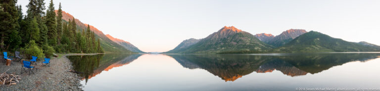 Blue camp chairs sit on a rocky shore beside a lake reflecting orange-lit mountains at dusk.