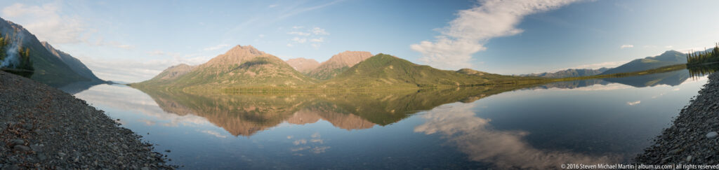 A wide mountain lake reflects pointed green and rocky peaks, with gravel shores in the foreground and clouds overhead.