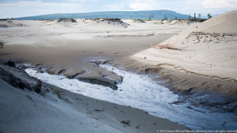 A narrow stream rushes through a pale sand cut and foams where it meets darker water.