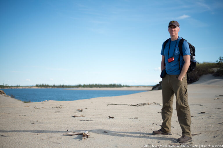 A man in a blue shirt and backpack stands on a broad sandy bank beside the river.