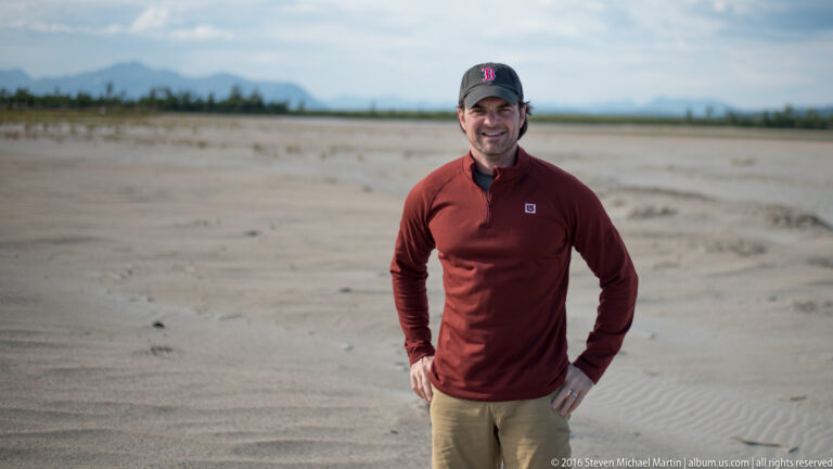 A man in a red shirt and dark cap stands on an open pale sand flat.