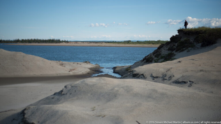 A small figure stands on a vegetated rise above a narrow channel between pale sandbanks and the river.