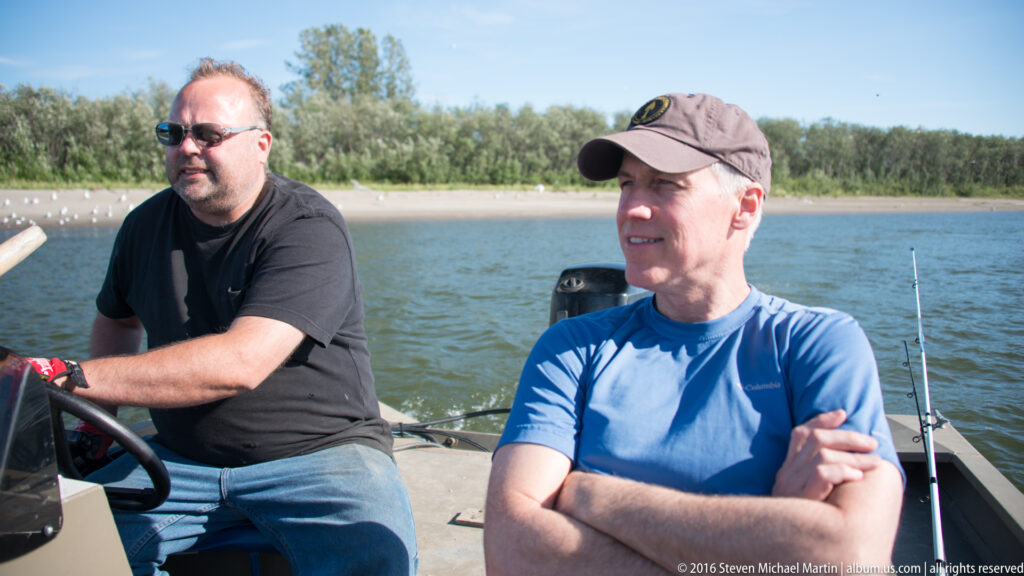Two men sit in a motorboat on the river, with an outboard motor and fishing rod behind them.