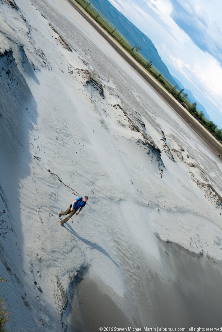 A small person in blue and red moves across a wide tilted view of a pale sandy bank.