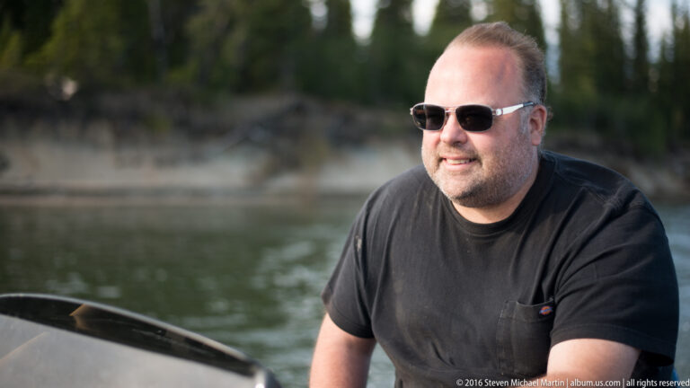 A man in dark sunglasses sits in a small boat with the river and shore behind him.