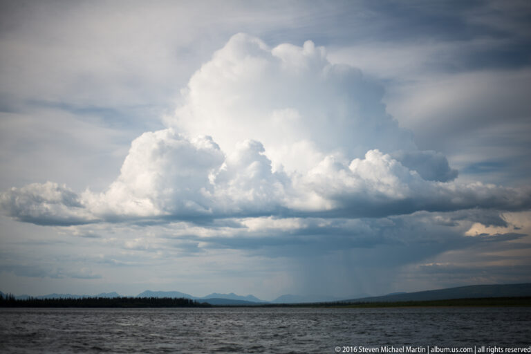 A large white cumulus cloud rises above a distant tree line and broad river.