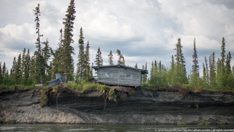 A weathered house stands at the top of a layered eroding bank above the river.