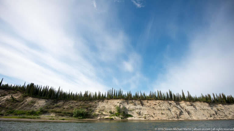 A pale bluff topped with spruce stretches above a narrow strip of river beneath streaked clouds.