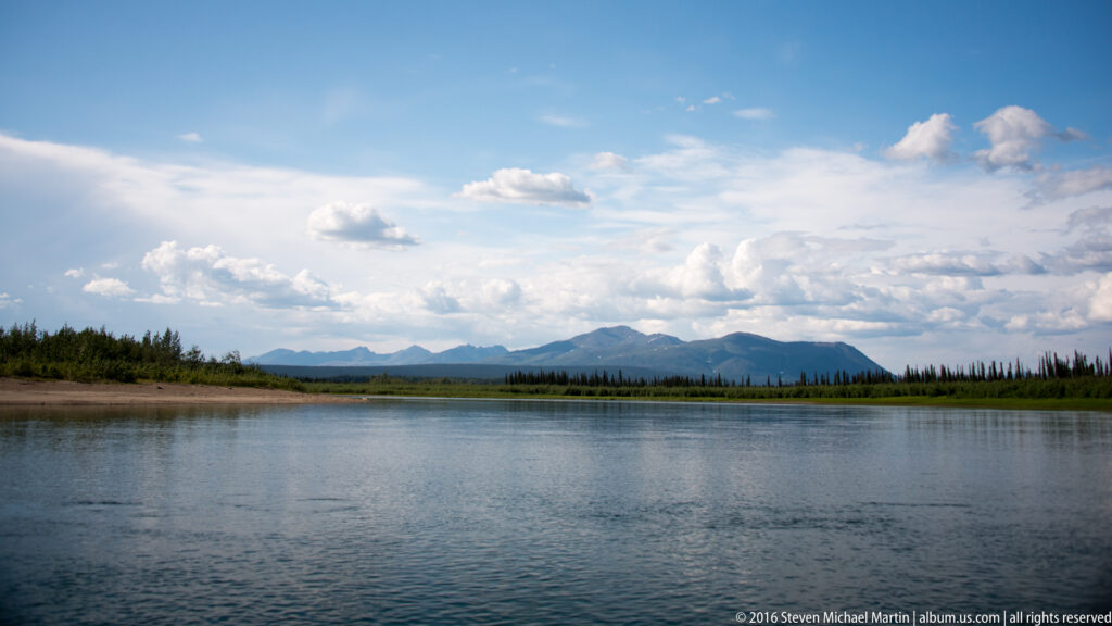 A broad river runs toward a spruce-lined bank and distant mountains beneath scattered clouds.