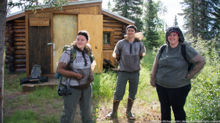 Three visitors in outdoor clothing stand beside a log cabin doorway.