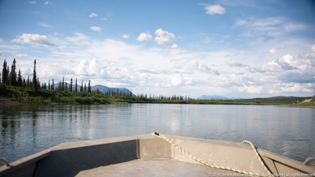 A boat bow and coiled rope point across open river water toward a low wooded shoreline.
